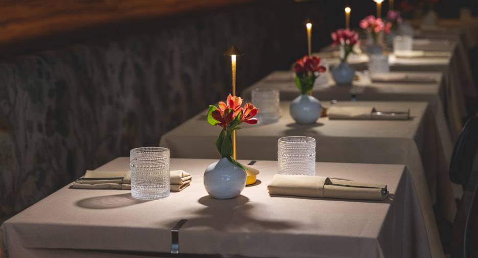 Tables set for two with beige tablecloths, glassware, napkins, small vases of red flowers, and lit candles in a dimly lit restaurant. Tables set for two with beige tablecloths, glassware, napkins, small vases of red flowers, and lit candles in a dimly lit restaurant.